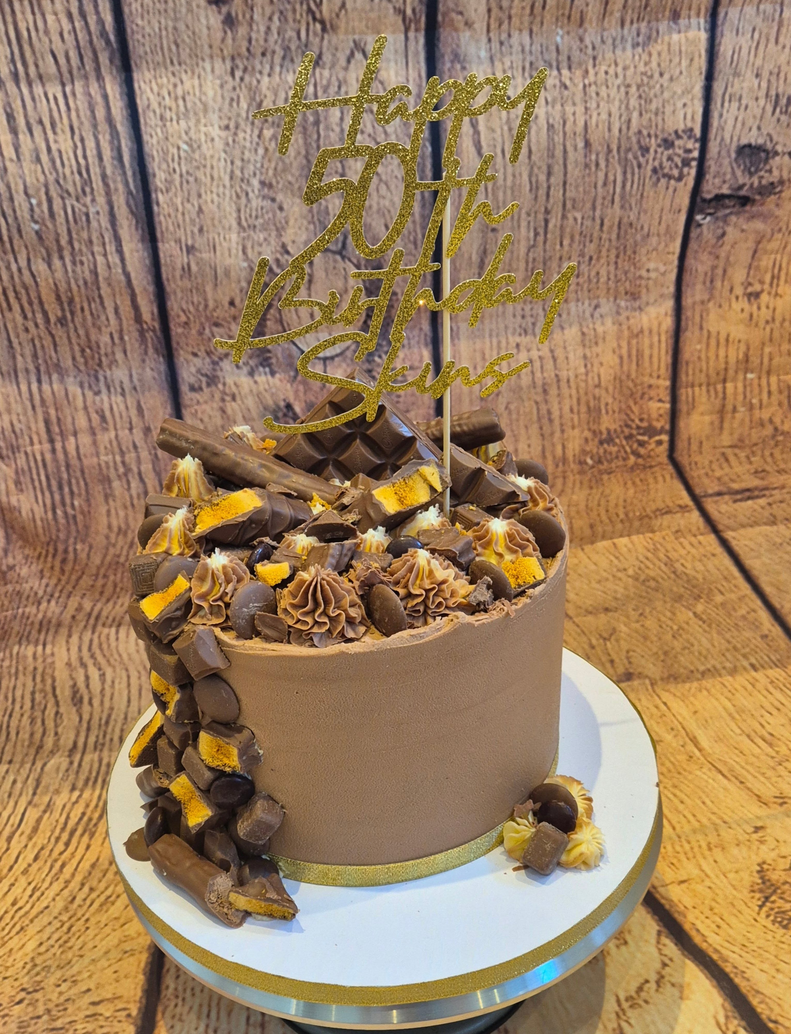 Rustic loose-iced chocolate celebration cake finished with smashed chocolate bars, gold sprays and a gold glitter '50th Birthday' topper